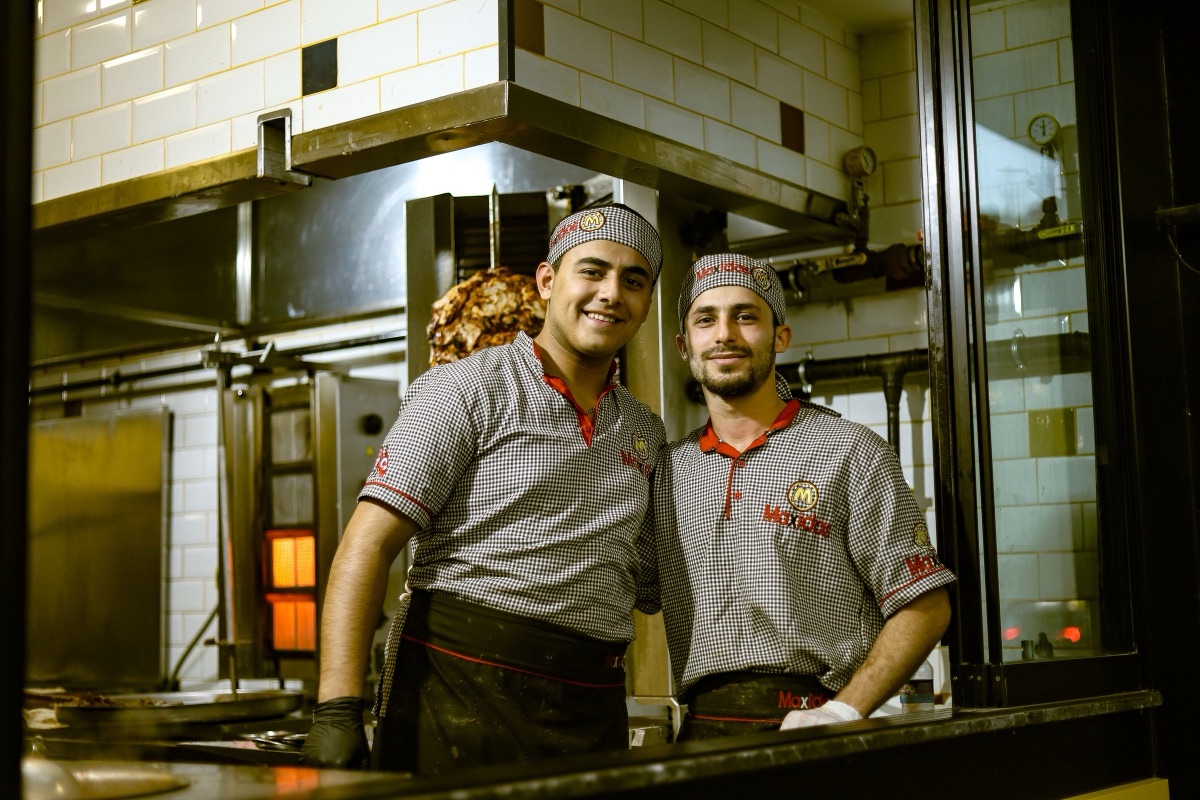 Two chefs in matching uniforms smiling in a busy kitchen